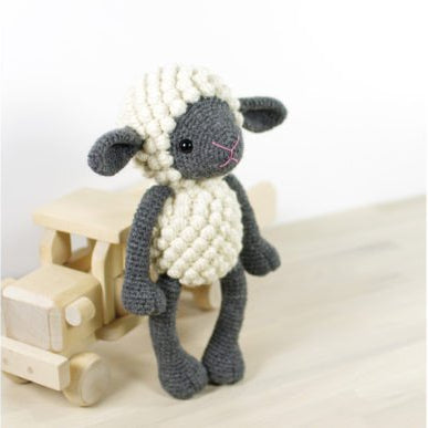 A crocheted plush sheep toy with a cream-colored wool body and dark gray face, legs, and ears, standing next to a small wooden toy truck on a light wood surface against a white background.